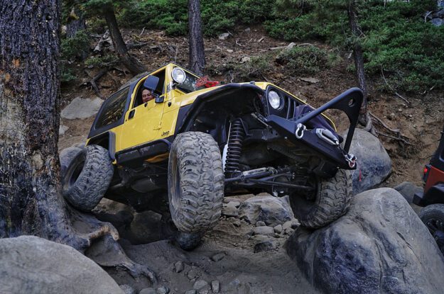 Yellow Jeep driving on rocks and roots with one wheel off the ground.